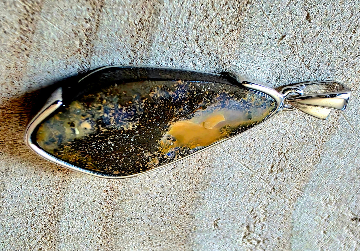A silver bezel-set pendant featuring a natural dark brown and black opal stone with a prominent golden-yellow inclusions, resting on a textured beige stone surface.
