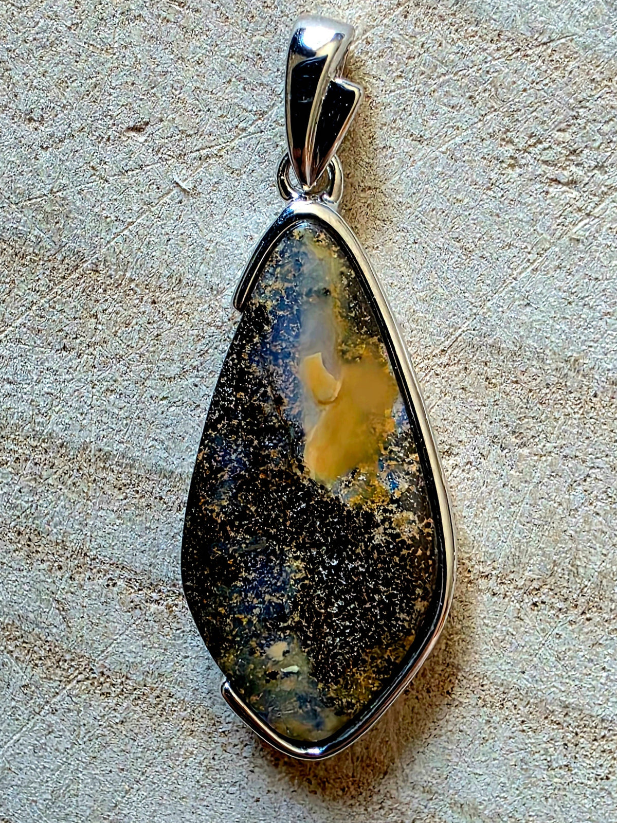 A teardrop-shaped pendant featuring a natural dark brown and black opal stone with a prominent golden-yellow inlay, set in a polished silver bezel with a silver bail, displayed on a textured beige surface.