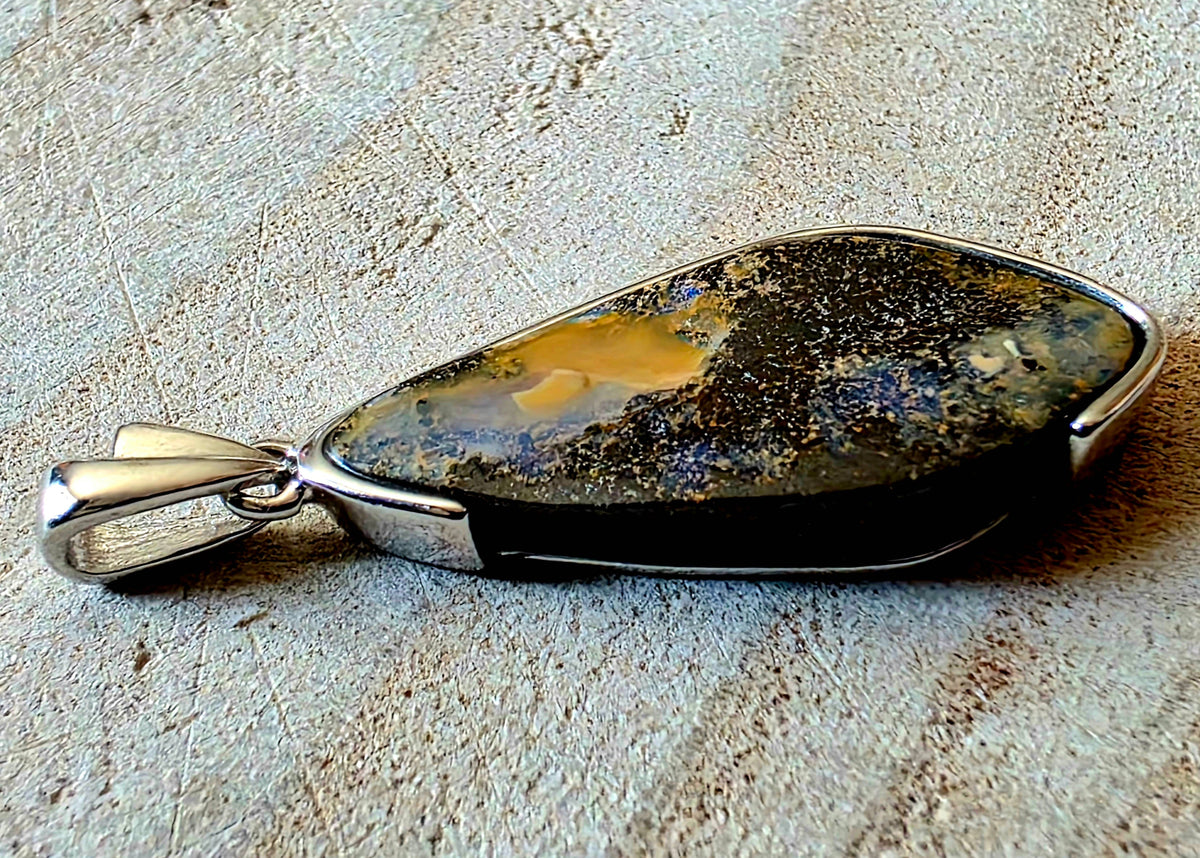 A teardrop-shaped pendant featuring a polished natural stone with a mix of black, gold, and blue hues, set in a polished silver-tone metal bezel with a bail, resting on a textured stone surface.