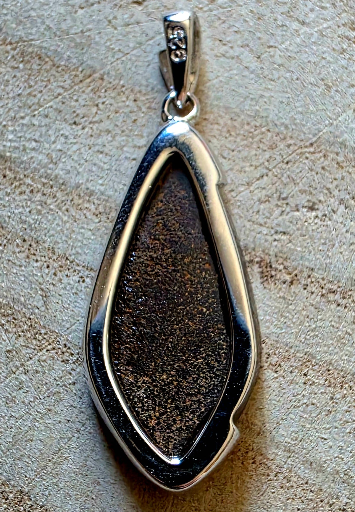 A silver teardrop pendant featuring a textured, glittery dark brown central opal, displayed on a neutral beige textured surface.