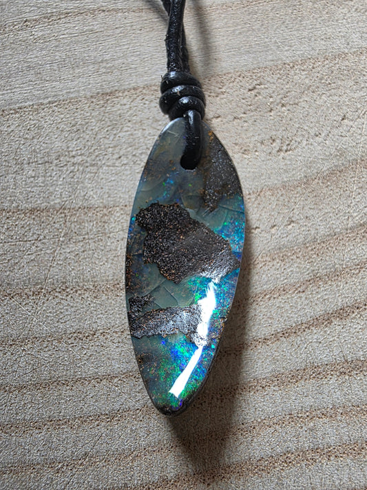 A teardrop-shaped lapel pendant made of polished iridescent opal with dark metallic inclusions, hanging from a black cord on a light wood background.