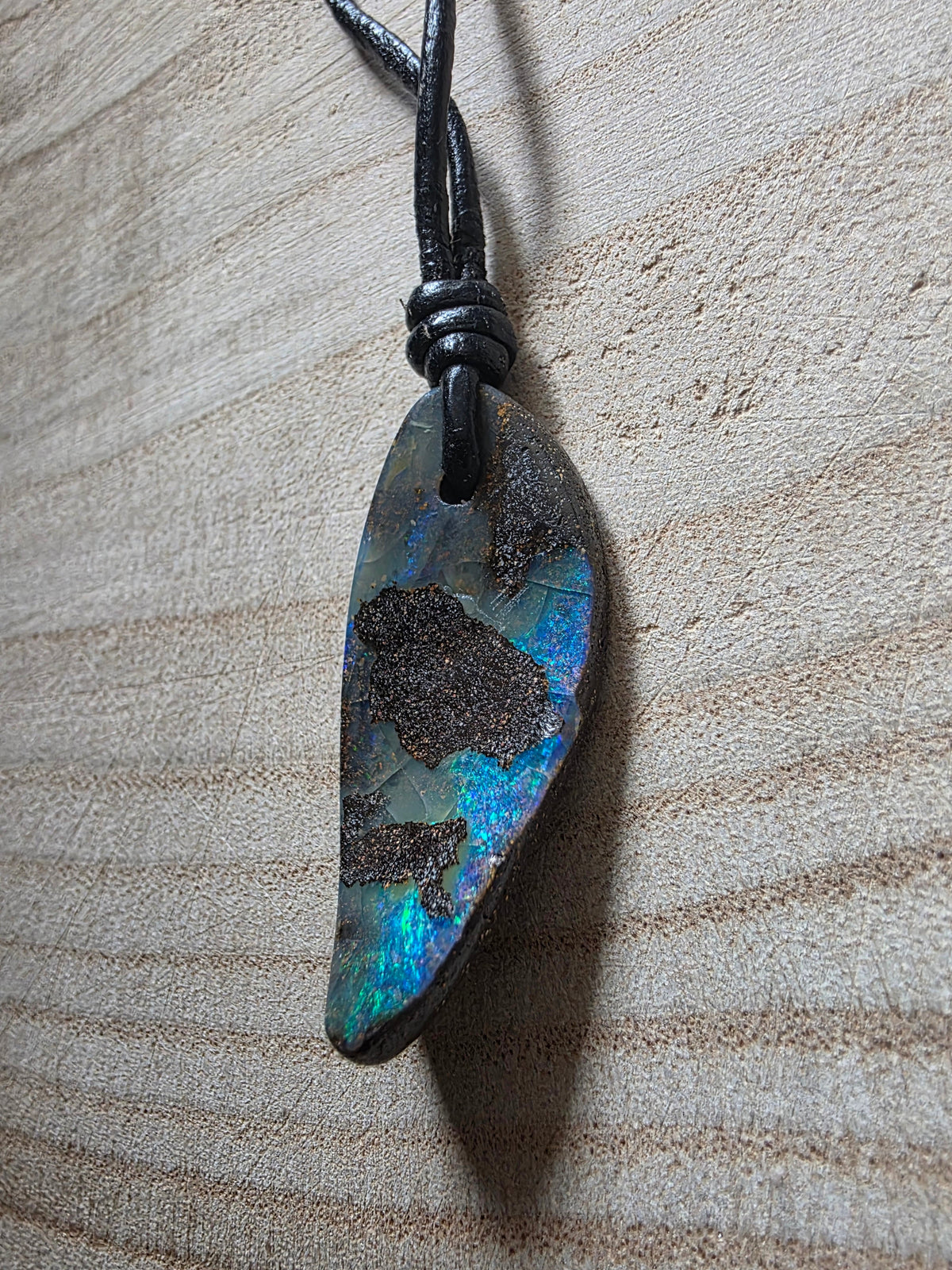A raw, irregular-shaped opal stone pendant with vibrant blue and green iridescence and dark matrix, suspended on a black cord against a light wood grain background.