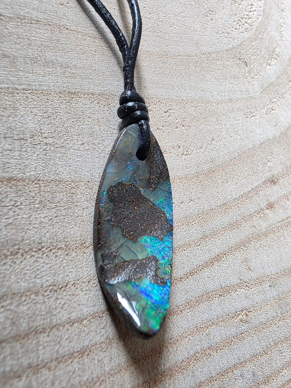A teardrop-shaped iridescent stone pendant with a black cord, resting on a light wood grain surface.