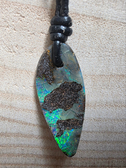 A teardrop-shaped pendant featuring a natural iridescent opal with dark inclusions, suspended from a black cord, set against a light wood grain background.