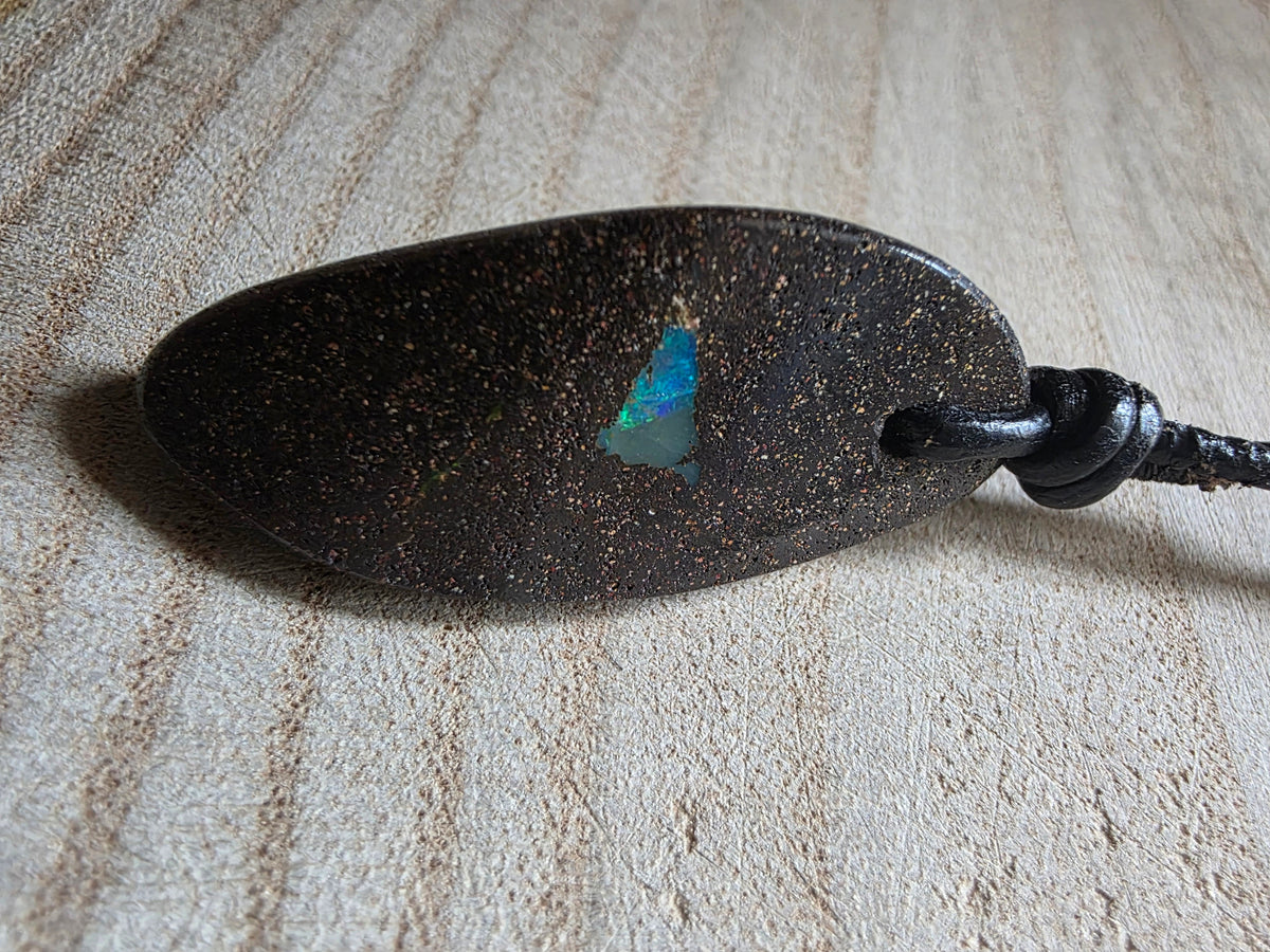 A dark brown speckled pendant with a small iridescent blue-green opal inlay, attached to a black cord, resting on a textured beige fabric background.
