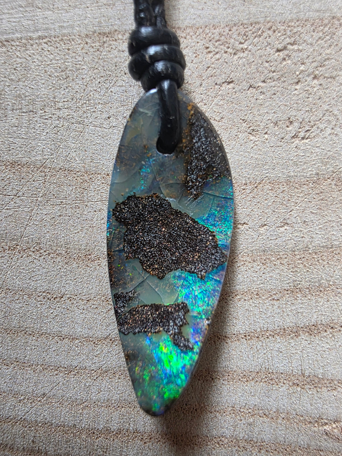 A teardrop-shaped pendant necklace featuring a natural iridescent opal stone with dark brown inclusions, suspended from a black braided cord on a textured beige background.