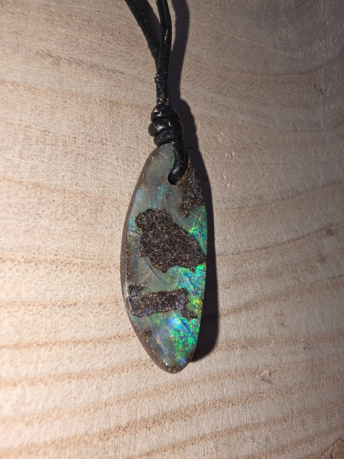 A teardrop-shaped iridescent opal pendant with dark inclusions, hanging from a black cord on a light wood background.