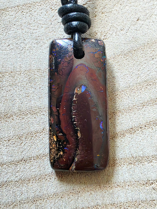 A polished rectangular opal pendant with swirling patterns of dark brown, black, and iridescent blue-gold, hanging from a black cord on a textured beige fabric background.