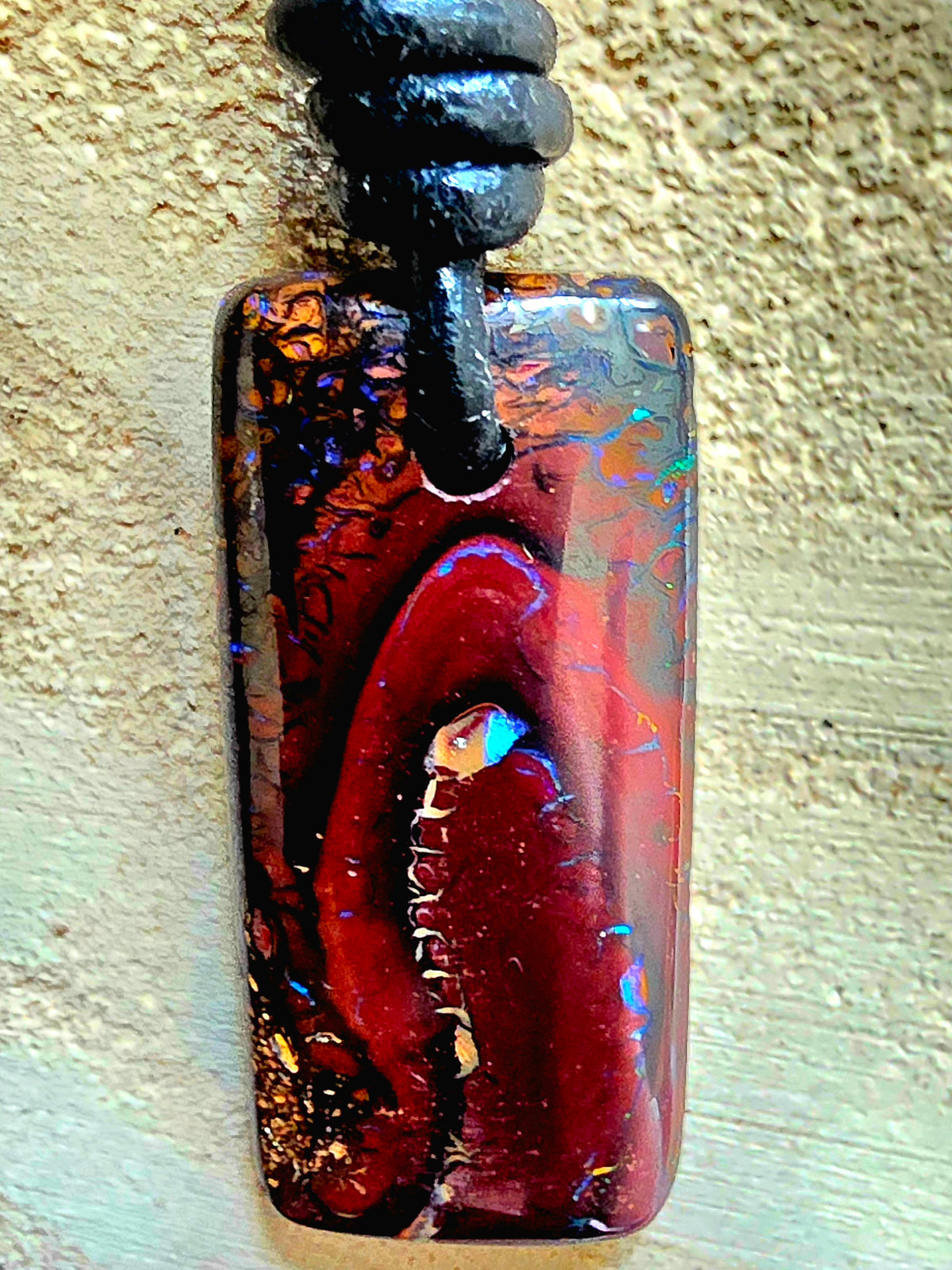 A rectangular pendant necklace featuring a polished brown black opal with iridescent blue and gold flecks, suspended from a black cord against a textured beige background.