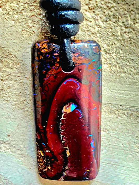 A rectangular pendant necklace featuring a polished brown black opal with iridescent blue and gold flecks, suspended from a black cord against a textured beige background.