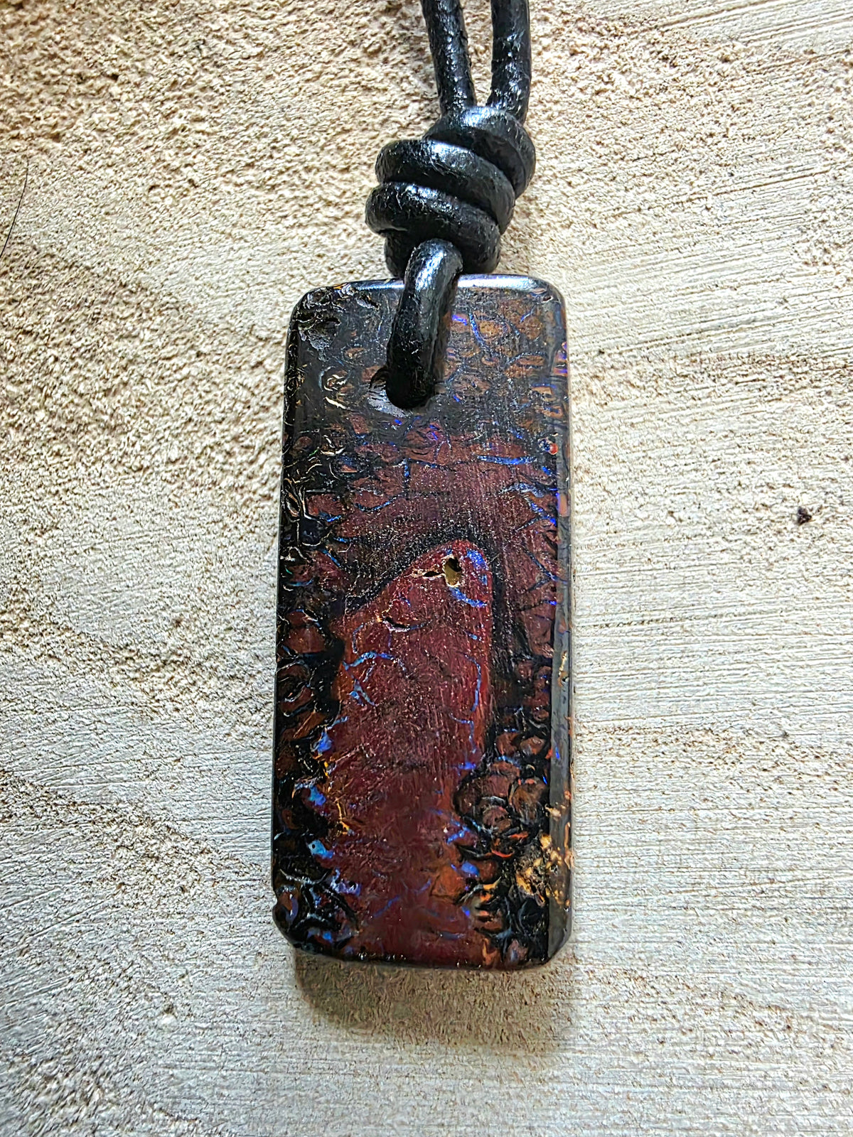 A rectangular boulder opal pendant necklace with a black cord, featuring a dark, iridescent surface with red, blue, and gold mineral-like patterns, resting on a textured beige surface.