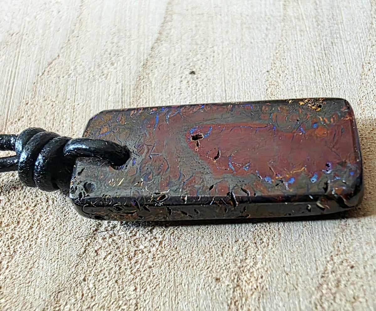 A rectangular iridescent opal stone pendant with a black cord, resting on a light beige textured surface. The stone displays a reddish-brown base with shimmering blue, purple, and gold hues.