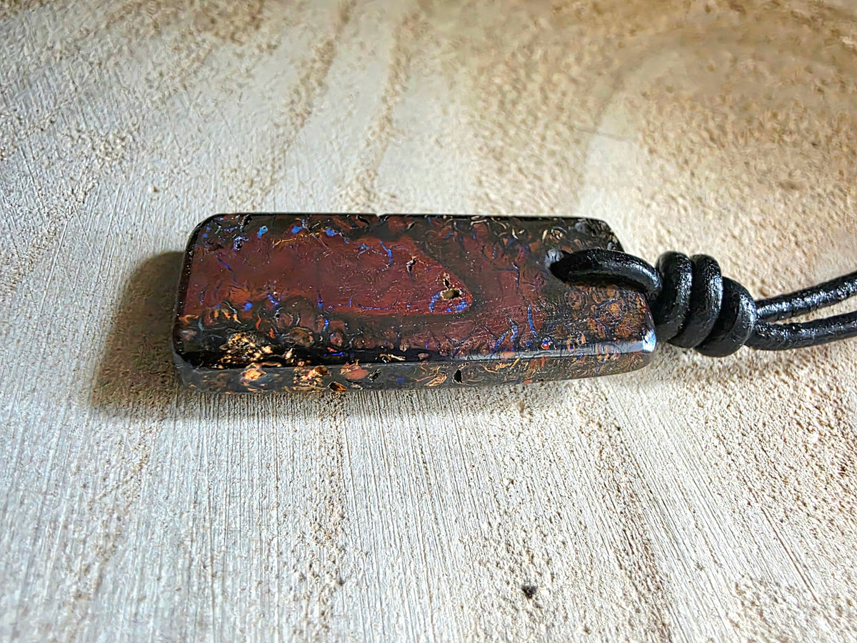 A polished rectangular boulder opal pendant with a dark reddish-brown iridescent surface and gold flecks, attached to a black cord, resting on a light wood surface.