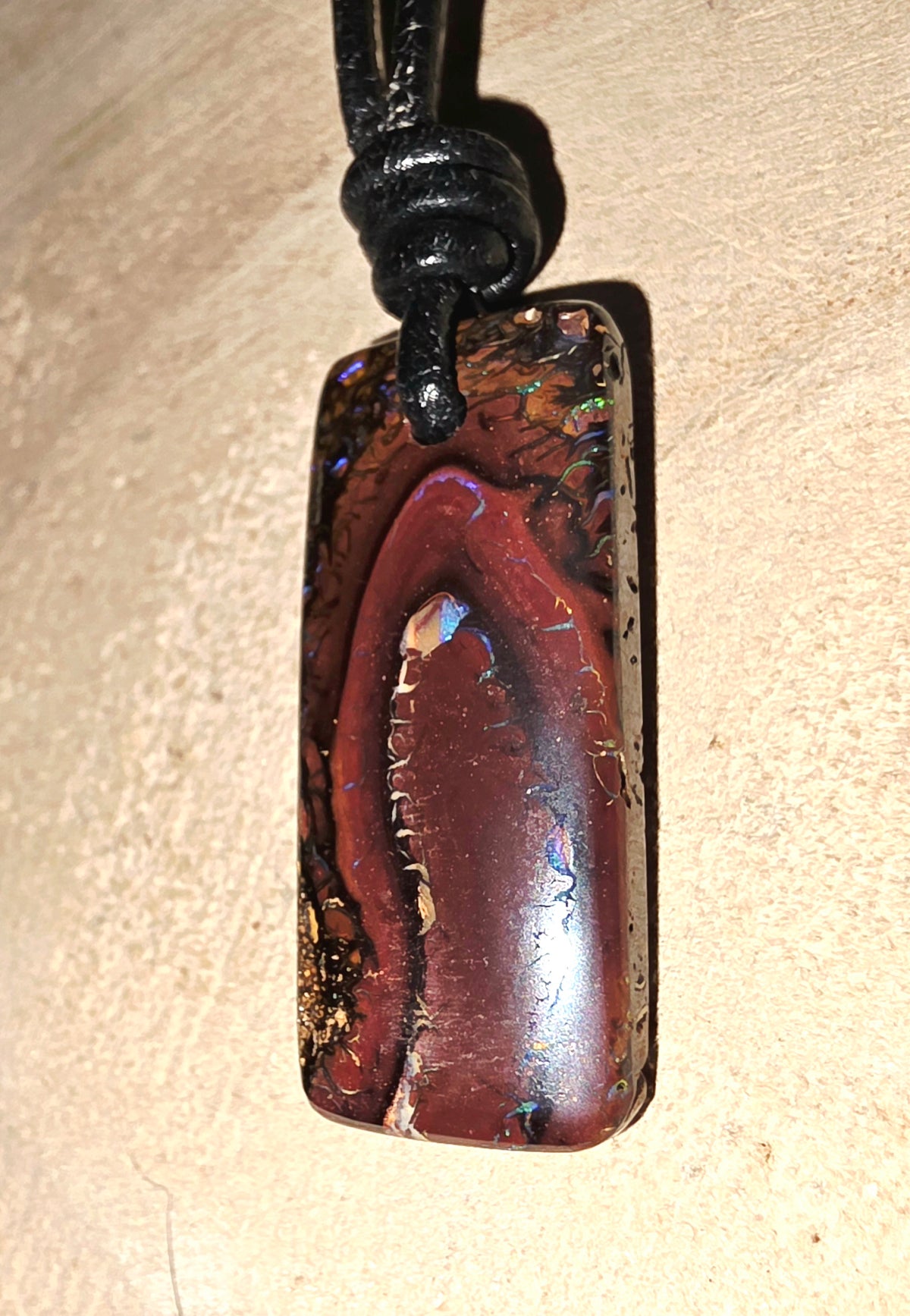 A rectangular pendant necklace made from a polished iridescent opal stone with a black cord, shown on a textured beige surface.