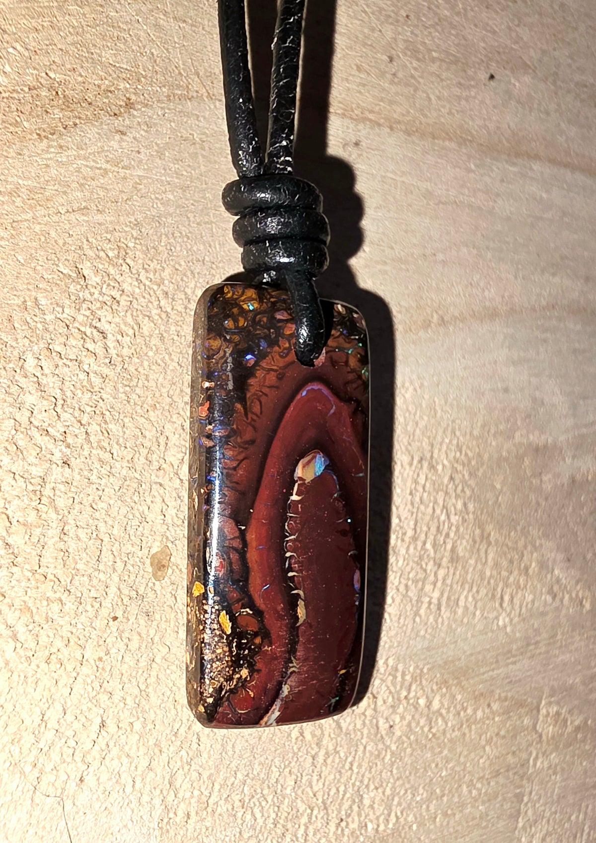 A rectangular pendant necklace with a black cord, featuring a polished opal stone with a deep red centre and multicoloured iridescent flecks, set against a light wood background.