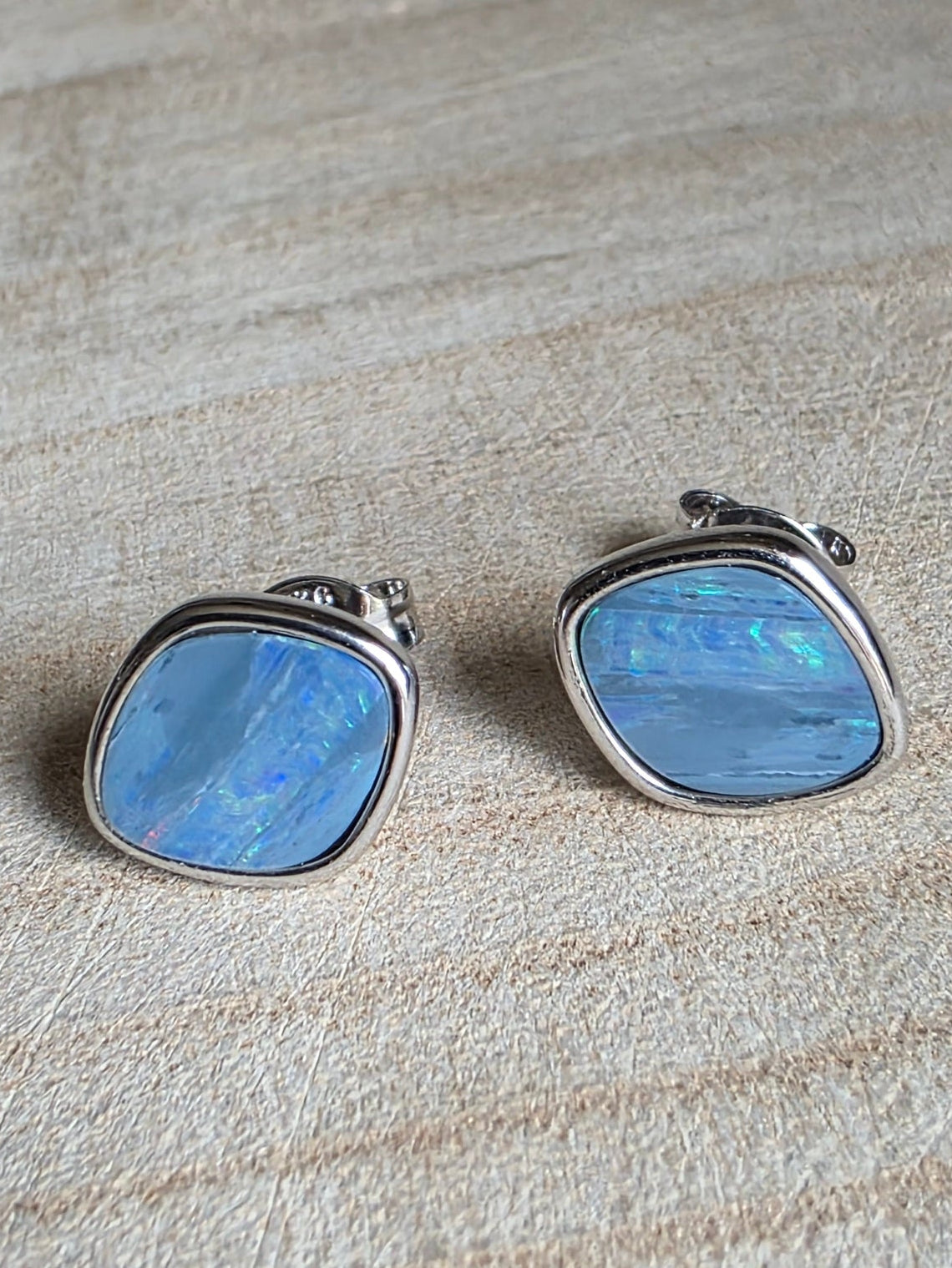 A pair of silver earrings with iridescent light blue square stones, resting on a light beige textured fabric surface.
