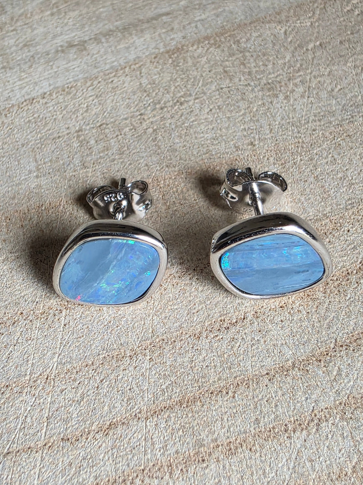 A pair of square stud earrings with iridescent blue-green stones in silver bezel settings, resting on a light wood surface.
