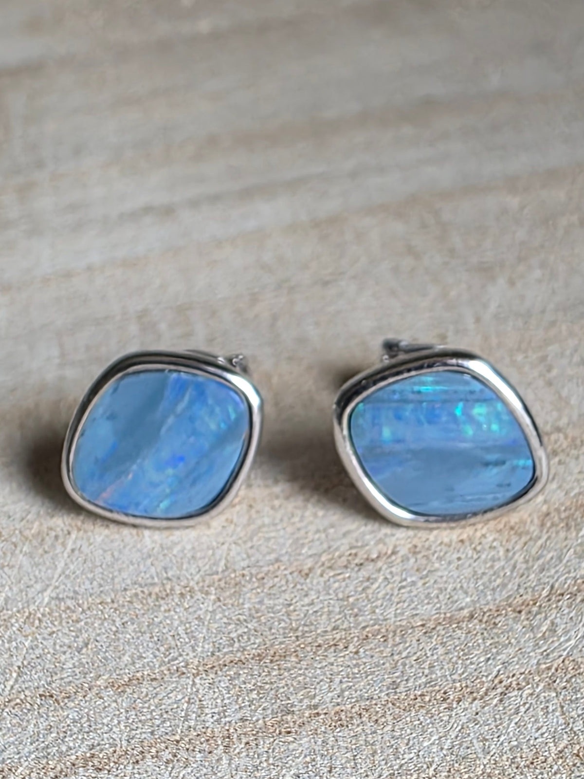 A pair of square stud earrings with iridescent light blue opal-like centres and silver bezels, resting on a light wood surface.