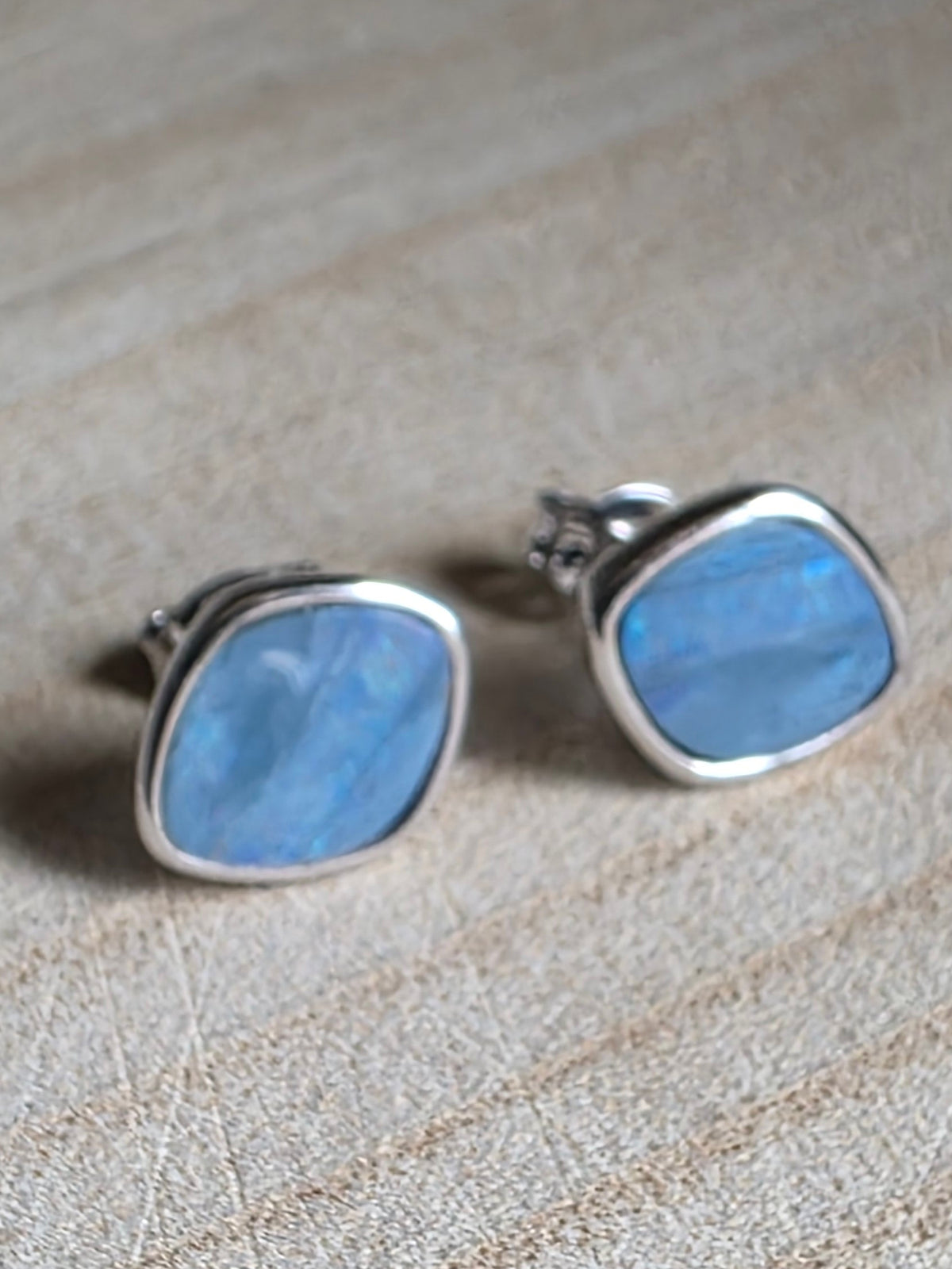 A pair of silver stud earrings with rounded, irregular blue opal gemstone inlays, resting on a textured beige fabric surface.