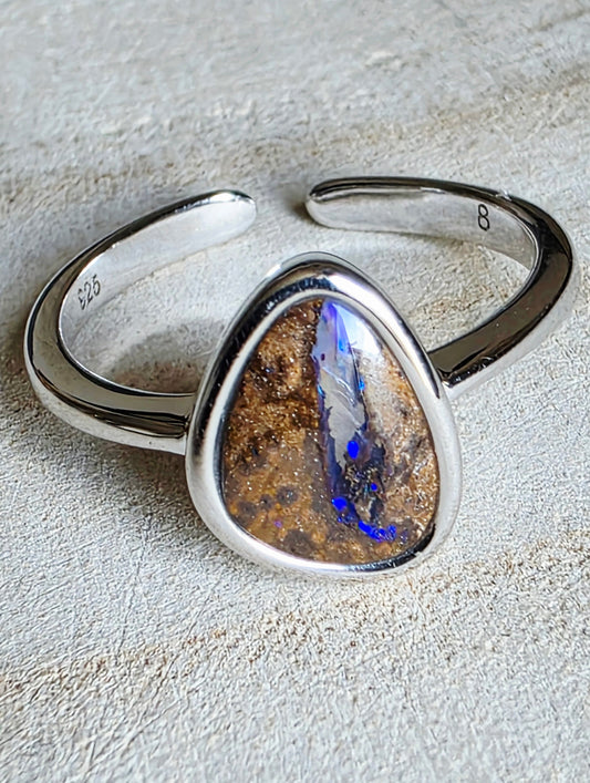 A sterling silver open cuff ring featuring a large, irregular triangular natural opal stone with brown and gold matrix and vibrant blue flashes, displayed on a textured light beige surface.