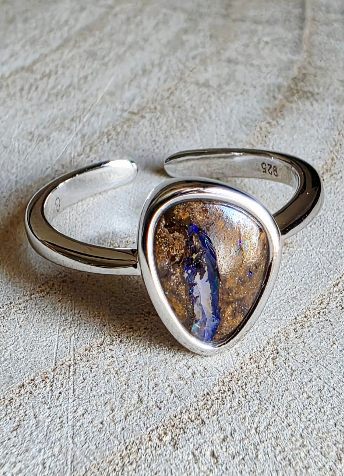 A sterling silver open band ring featuring a large, irregular teardrop-shaped cabochon opal stone with a brown, gold, and blue iridescent pattern, resting on a textured beige surface.