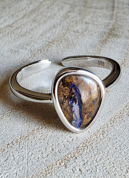 A sterling silver open band ring featuring a large, irregular teardrop-shaped cabochon opal stone with a brown, gold, and blue iridescent pattern, resting on a textured beige surface.