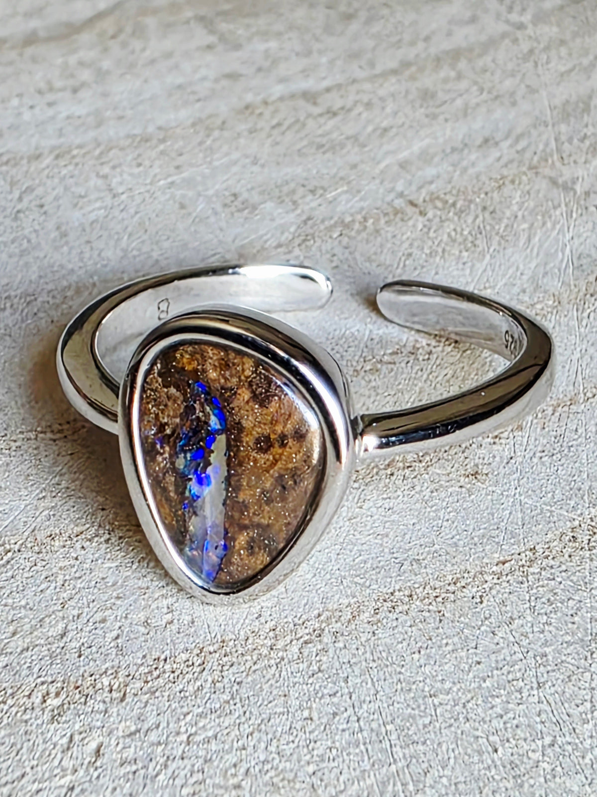 A silver open cuff ring featuring a large, irregular teardrop-shaped opal stone with brown matrix and vivid blue flashes, set on a textured light beige surface.