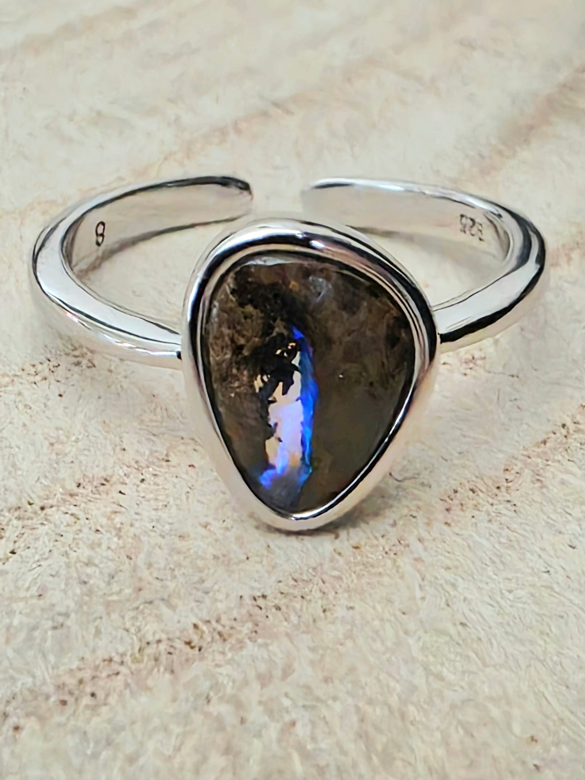 A sterling silver open shank ring featuring a teardrop-shaped labradorite stone with a blue and orange flash, resting on a textured beige surface.