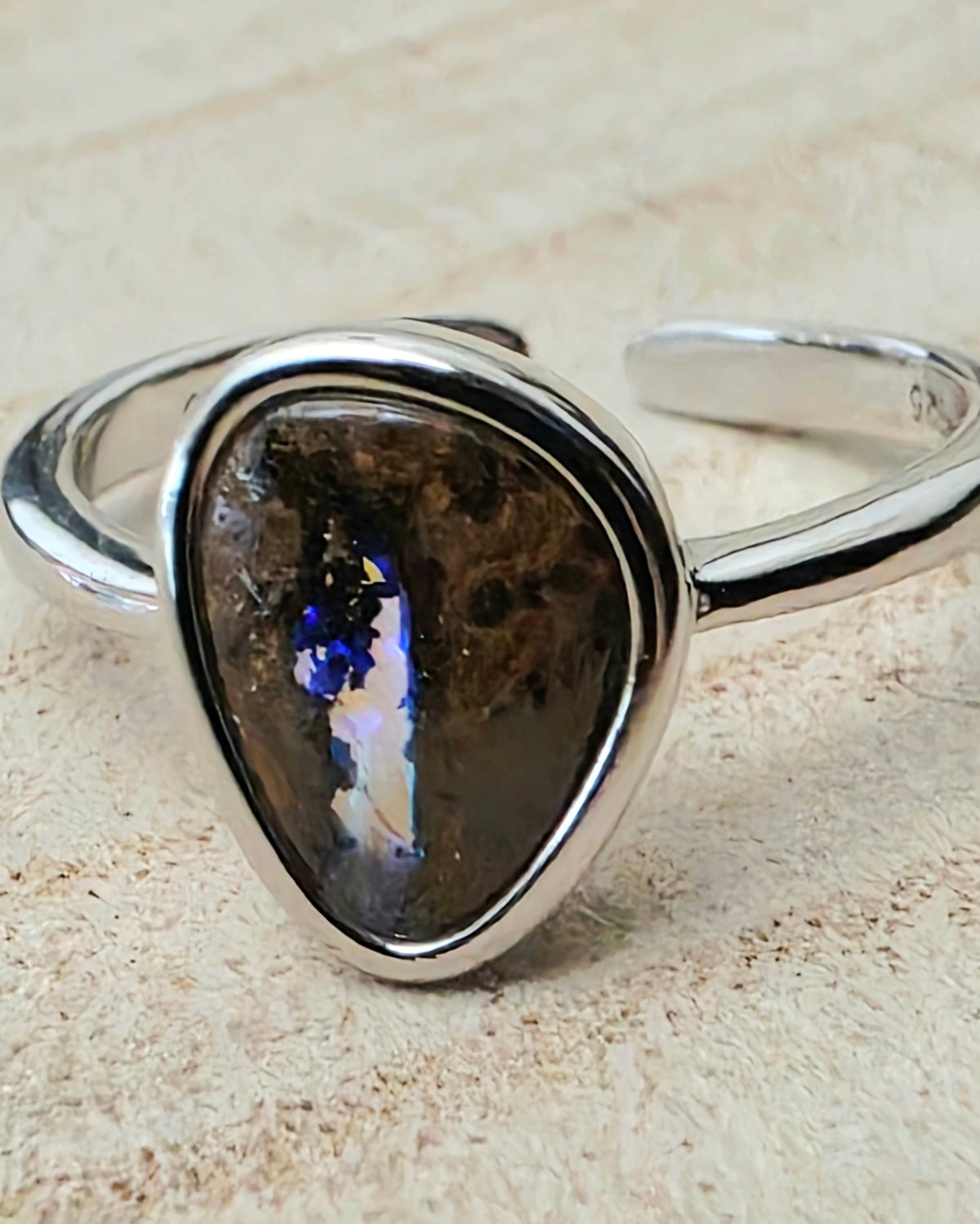 A silver-toned open band ring featuring a large, irregular teardrop-shaped dark brown p[al stone with vibrant blue and white iridescent flashes, resting on a textured beige surface.