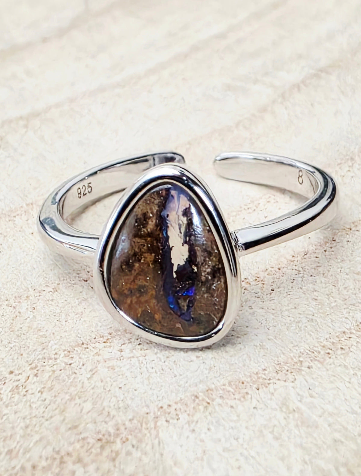 A sterling silver open band ring featuring a large, irregular triangular bezel-set opal stone with a brown and black mottled pattern and a vibrant blue flash, displayed on a textured beige surface.