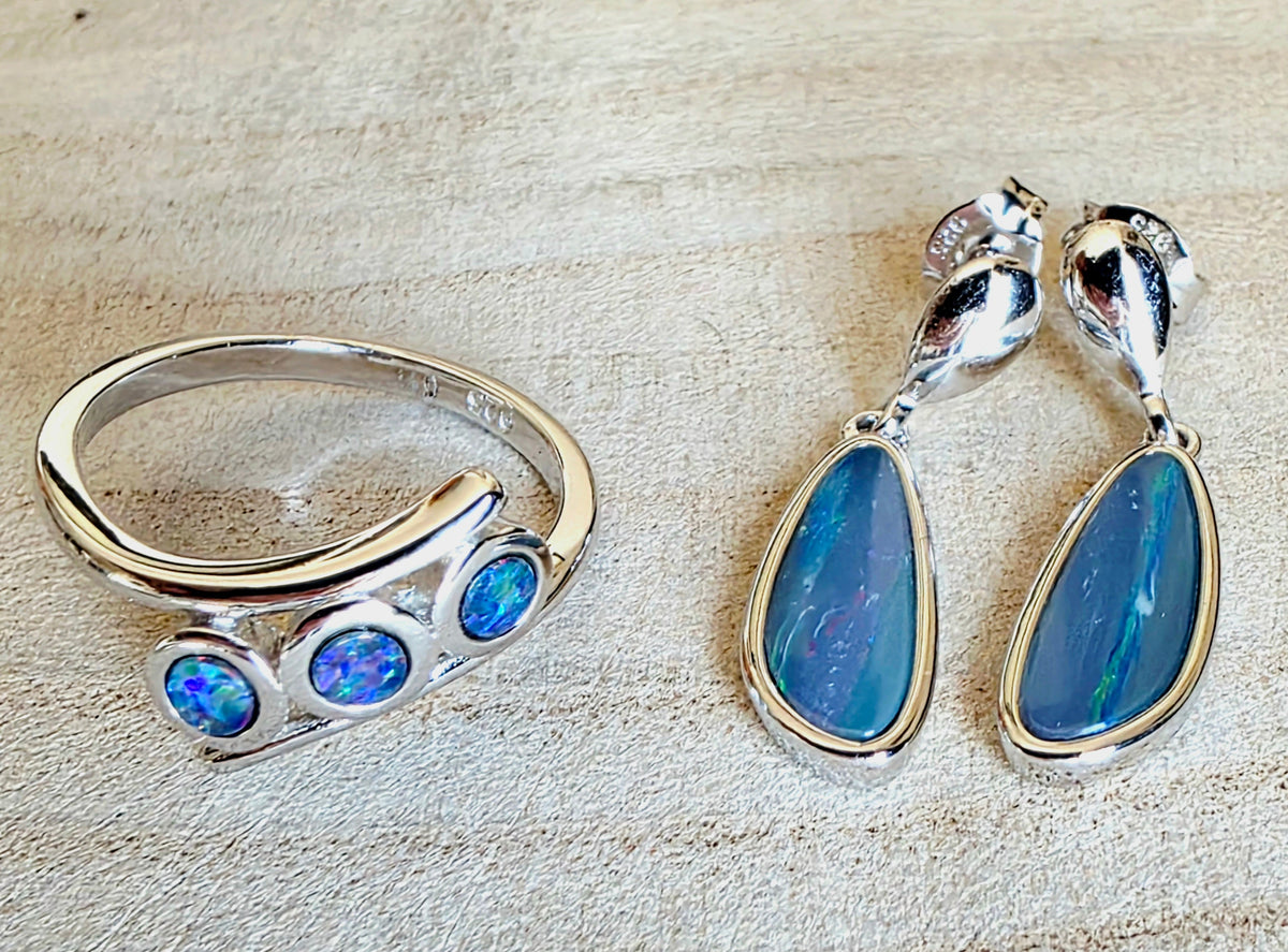 A sterling silver jewellery set featuring a curved ring with three round blue opal stones and matching drop earrings with teardrop-shaped blue opal stones, displayed on a textured beige surface.