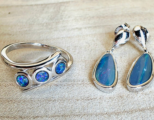 A sterling silver ring and matching drop earrings featuring vibrant blue opal gemstones, displayed on a light beige textured background.