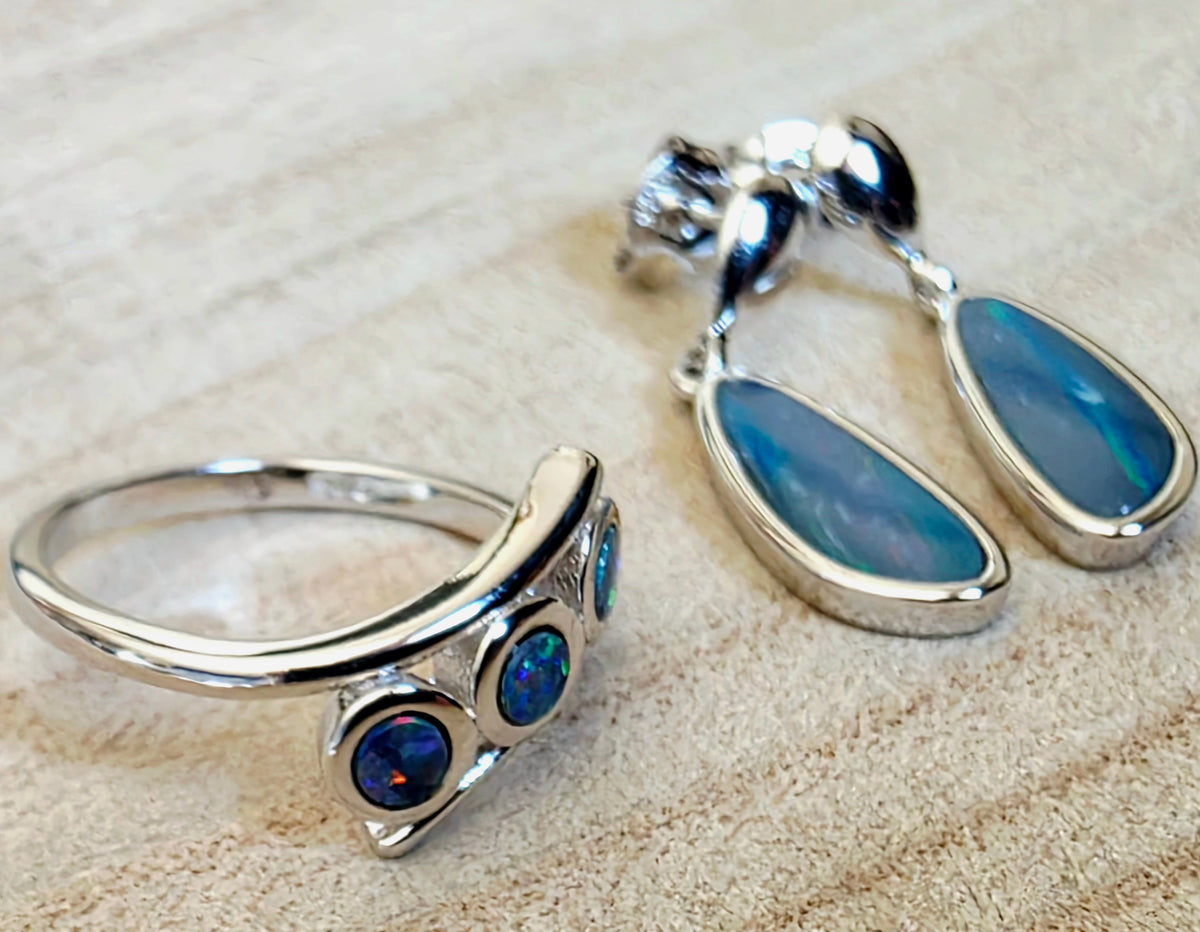 A sterling silver ring and matching drop earrings with blue opal stones, displayed on a textured beige background.