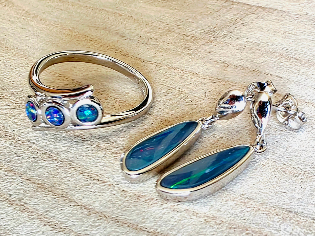 A sterling silver ring and matching drop earrings featuring blue opal gemstones, displayed on a light wood surface.