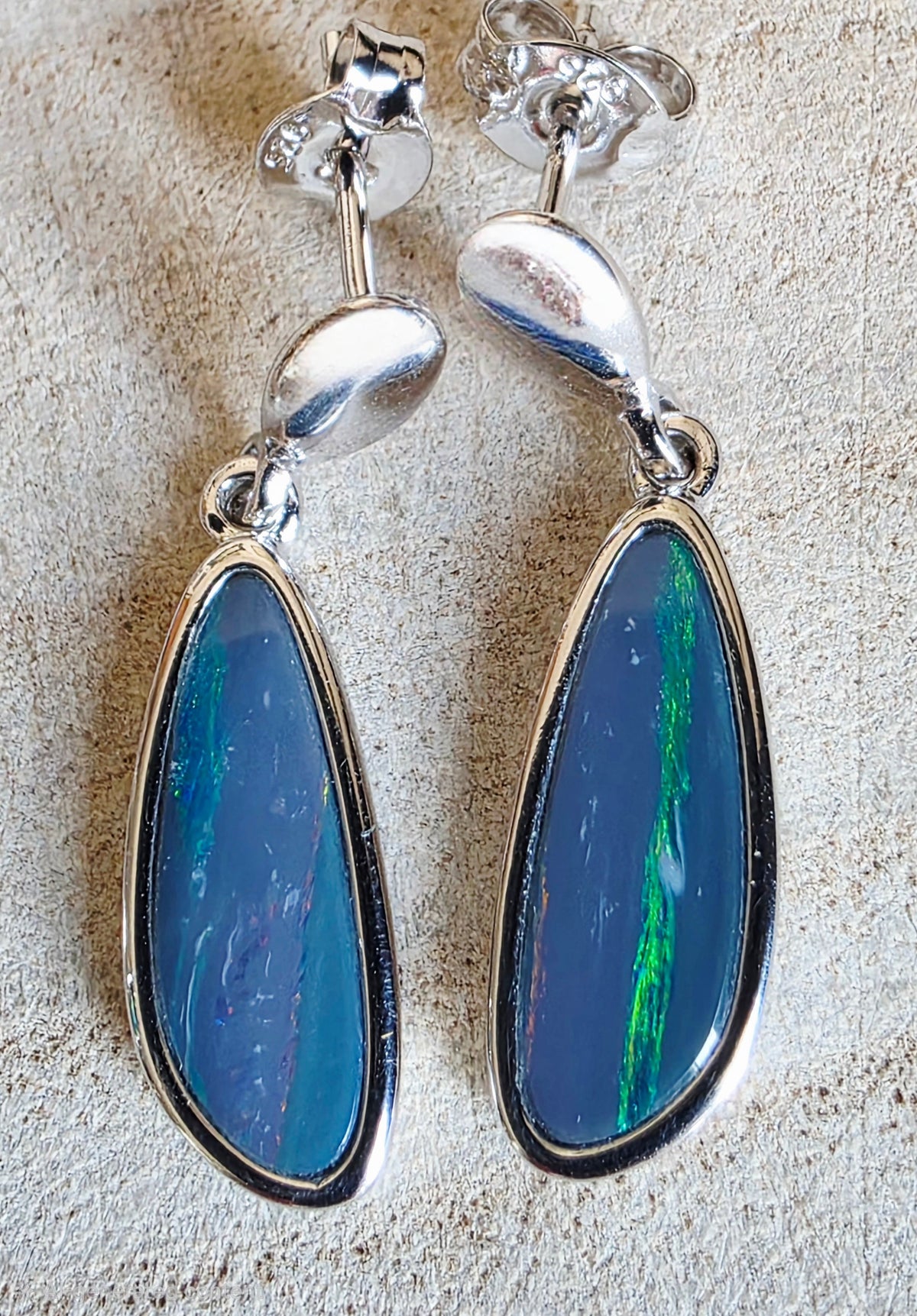 A pair of silver dangle earrings with teardrop-shaped blue opal stones, displayed on a textured beige surface.