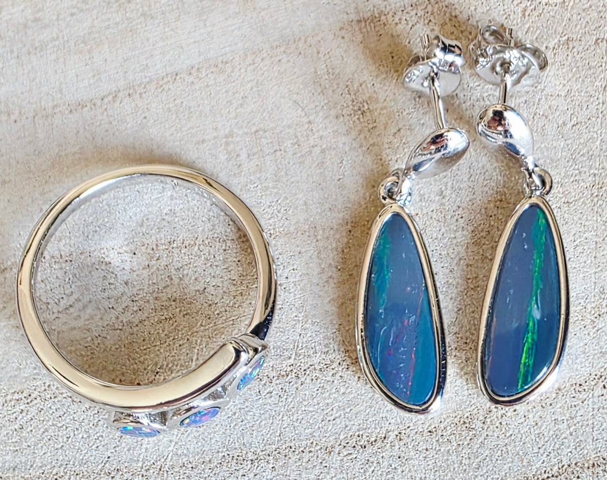 A sterling silver jewellery set featuring a ring and matching drop earrings with teardrop-shaped blue opal inlays, displayed on a textured beige background.