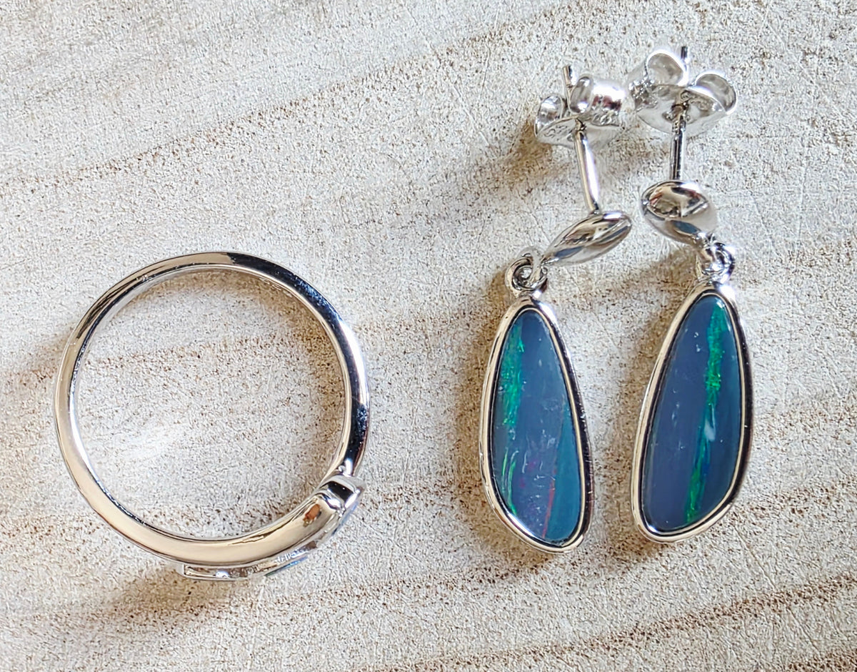 A sterling silver jewellery set featuring a an opal ring and a pair of drop earrings with teardrop-shaped blue opal stones, displayed on a textured beige surface.