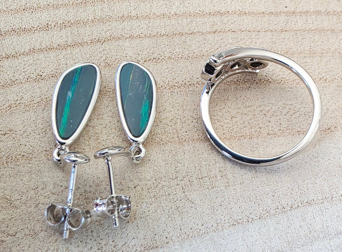 A pair of silver drop earrings with teardrop-shaped green opal inlays and matching silver ring, displayed on a textured beige background.