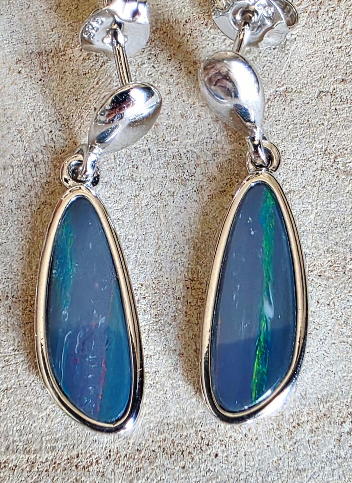 A pair of silver dangle earrings with teardrop-shaped blue iridescent opal stones in bezel settings, displayed on a textured beige surface.