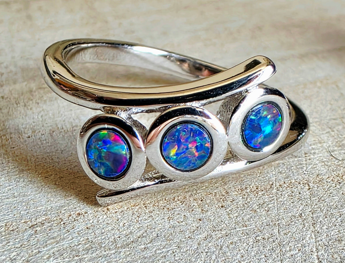 A sterling silver ring featuring three oval-cut blue and multicoloured opal gemstones, displayed on a textured beige surface.
