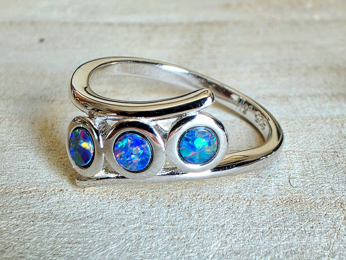 A sterling silver ring featuring three round blue opal gemstones in bezel settings, displayed on a textured beige background.