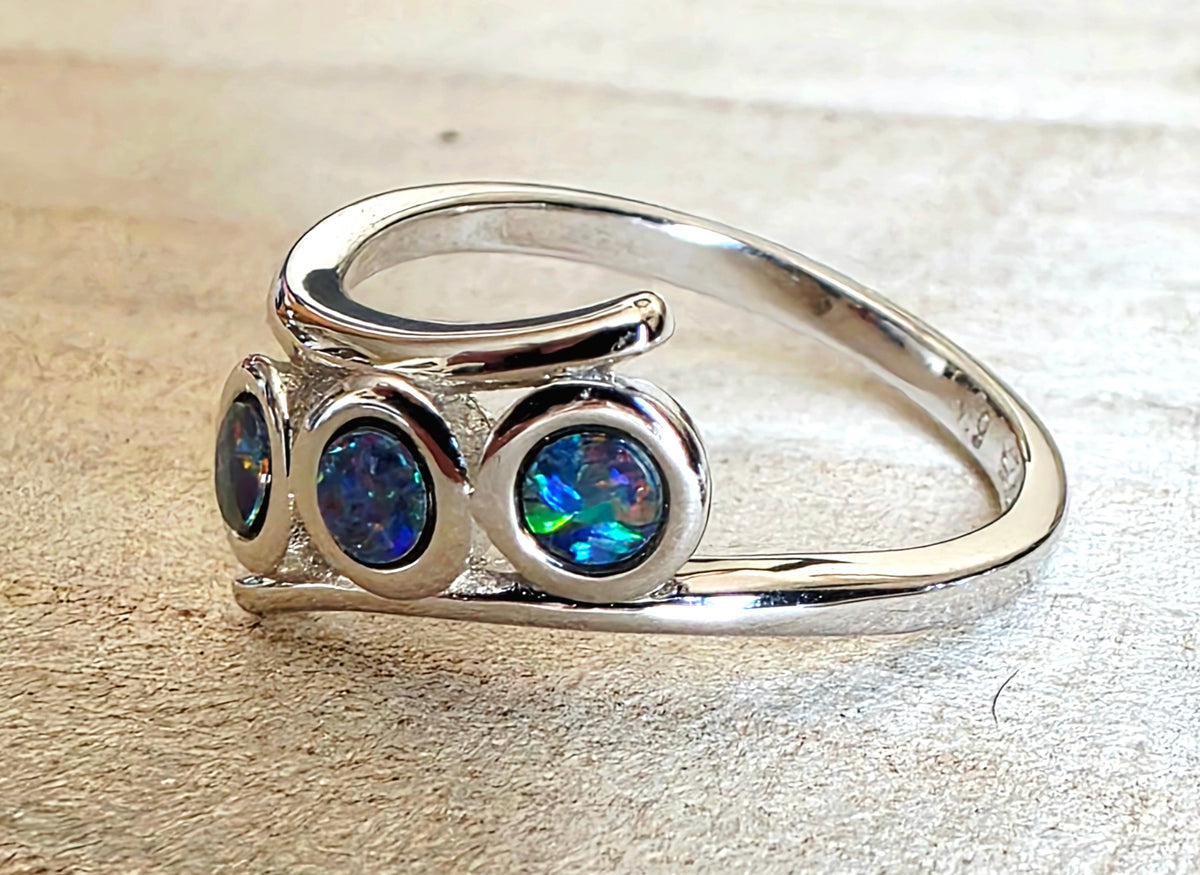 A polished silver ring featuring three round iridescent blue opal gemstones in bezel settings, displayed on a textured beige surface.