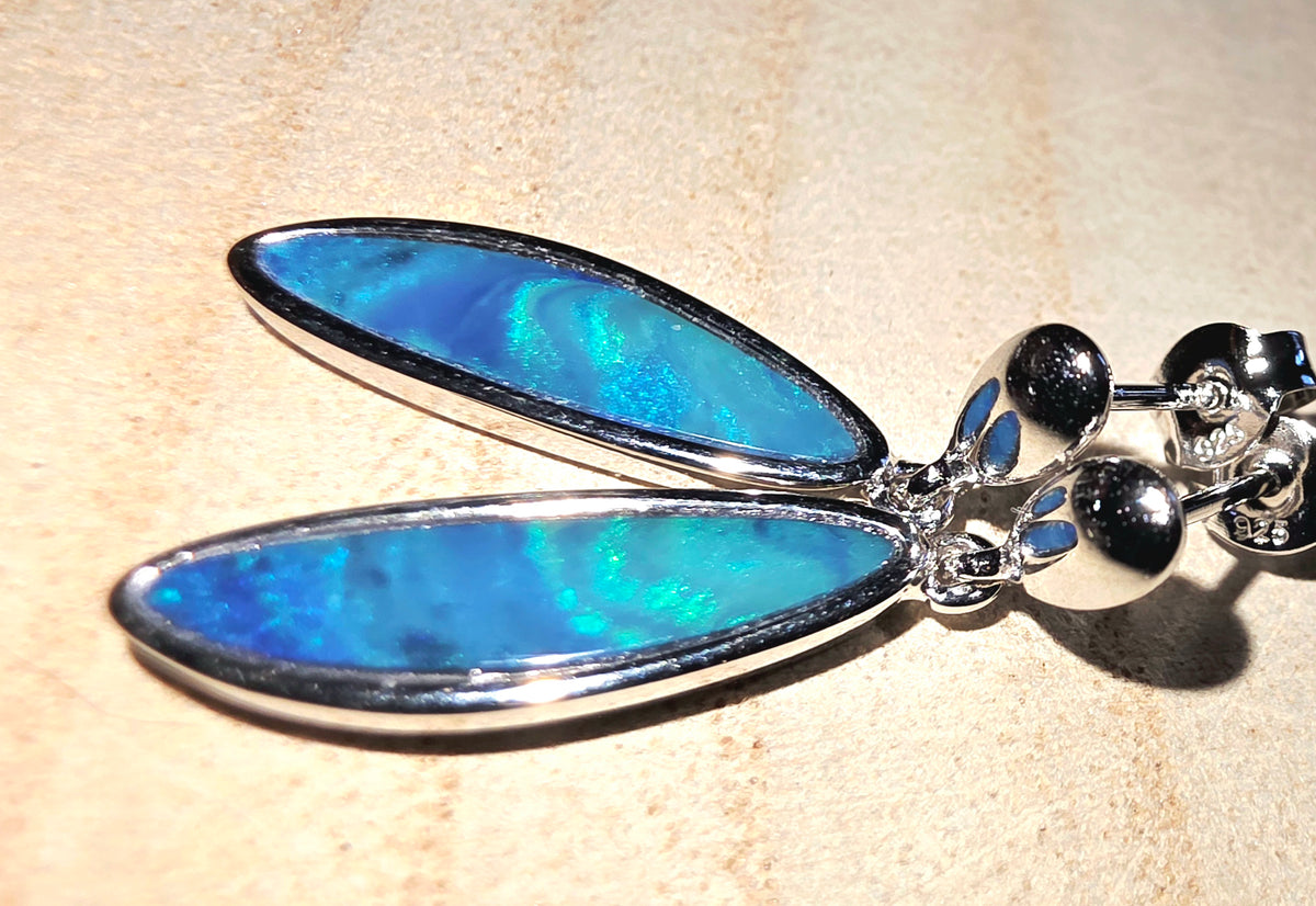A close-up of a silver earring featuring two elongated oval opals with a vivid blue iridescent finish, set against a light beige textured background.