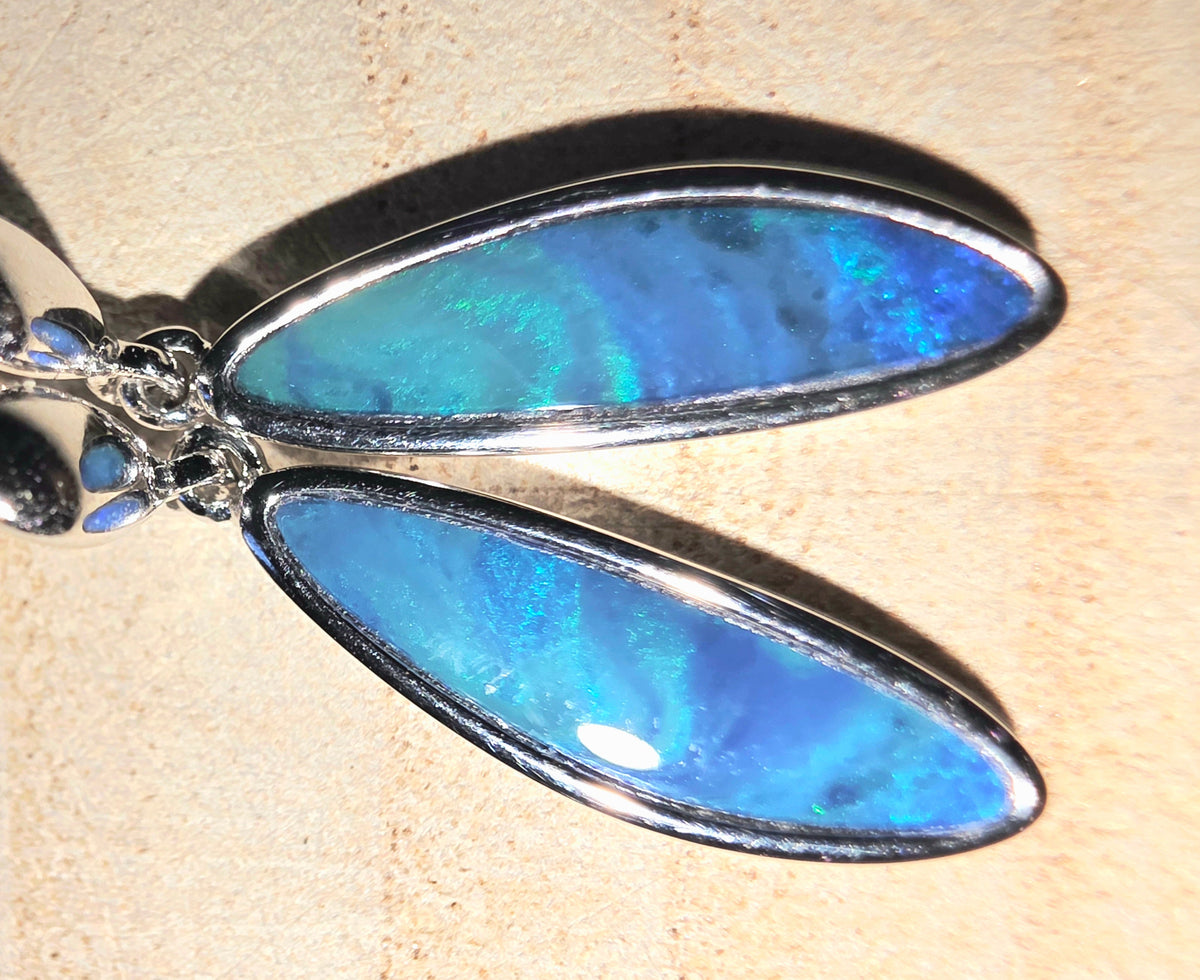 A close-up of a pair of silver dangle earrings with teardrop-shaped blue iridescent opal stones, resting on a textured beige surface.