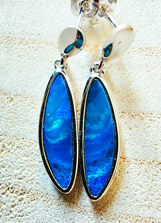 A pair of silver drop earrings featuring large, teardrop-shaped blue iridescent opal stones, displayed on a textured beige surface.