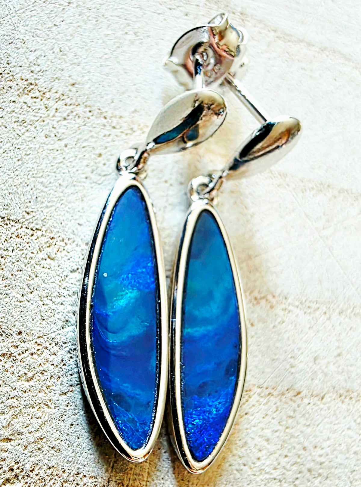A pair of silver drop earrings with teardrop-shaped blue iridescent opal inlays, displayed on a textured beige surface.