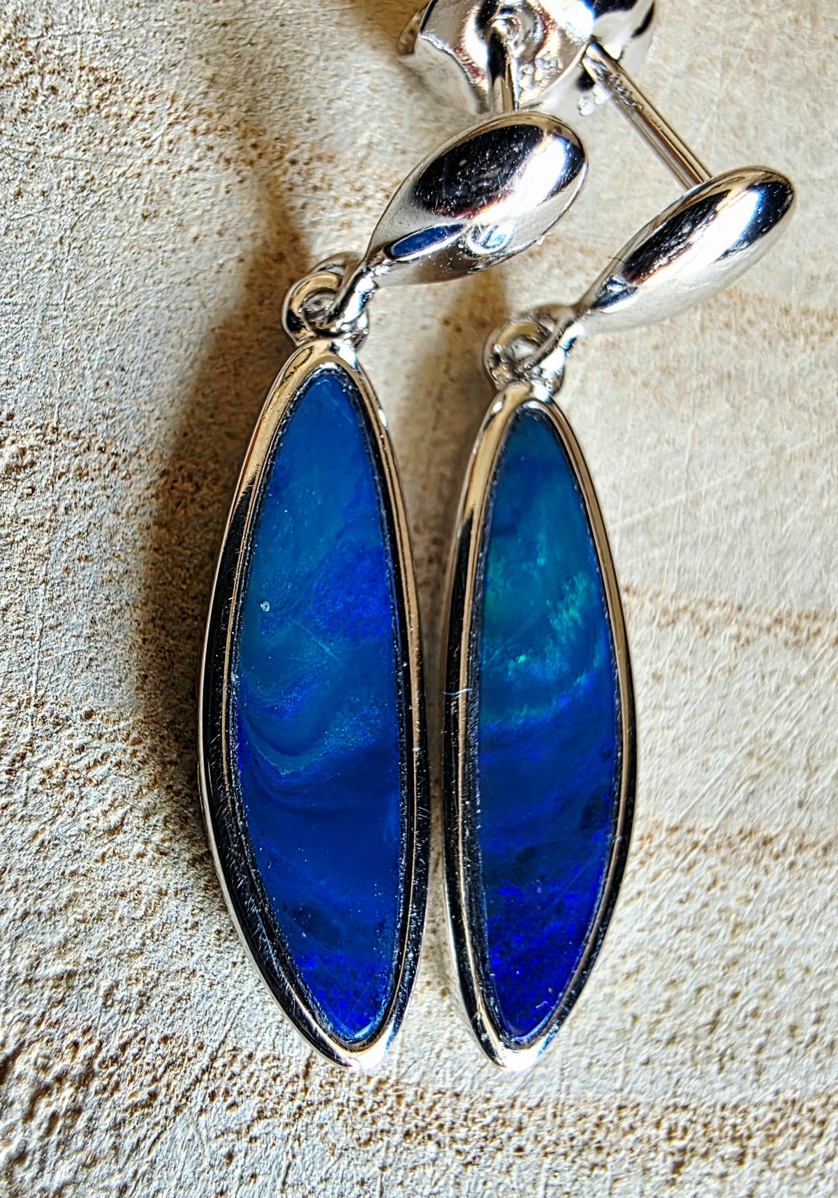 A pair of silver drop earrings with marquise-shaped blue opal inlays, displayed on a textured beige surface.