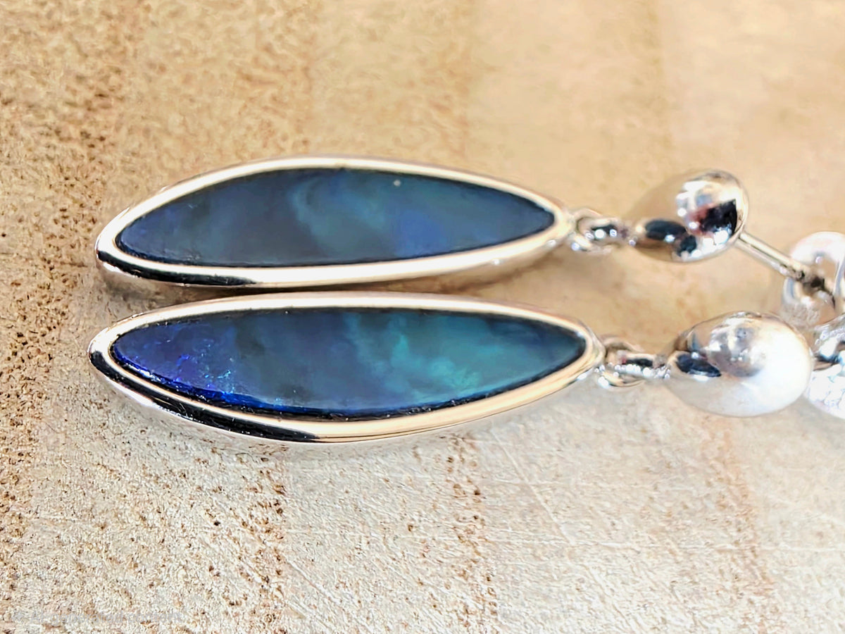 A pair of silver dangle earrings with teardrop-shaped blue iridescent opal gemstones and small silver beads, displayed on a textured beige surface.