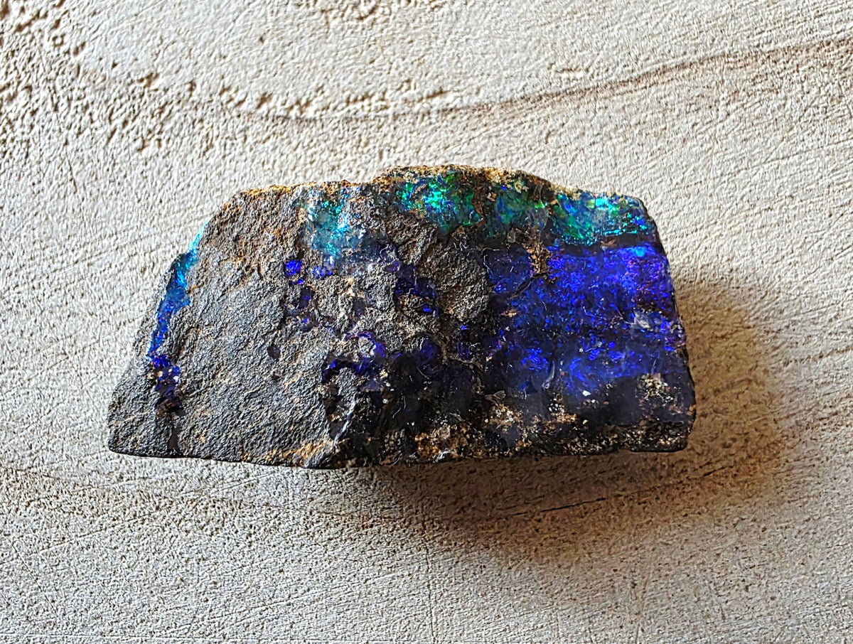 A rough natural opal stone with vibrant blue, teal, and purple iridescent inclusions on a dark matrix, photographed on a textured beige surface.