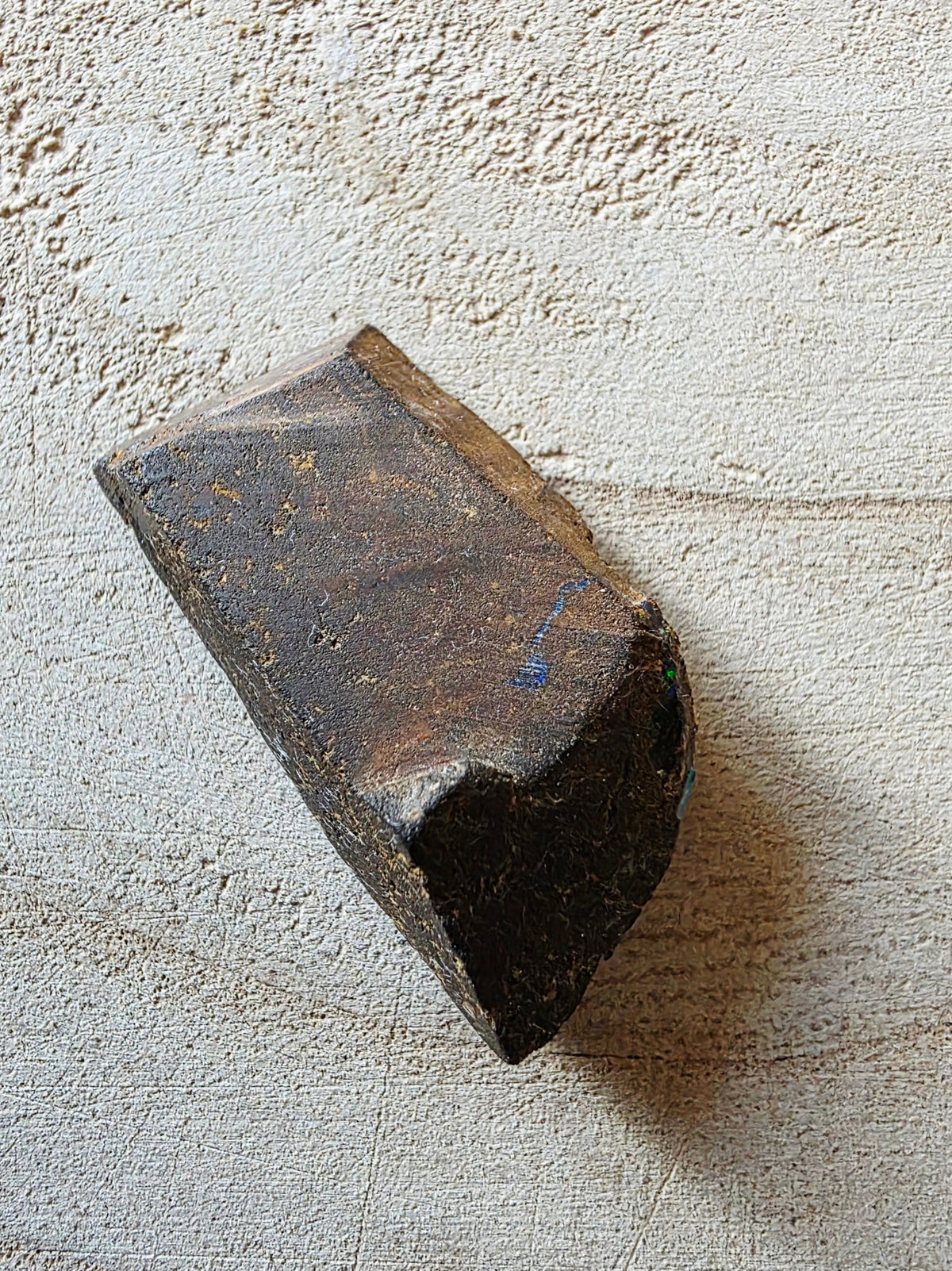A dark brown, rectangular opal with a rough, weathered texture and a chipped edge, resting on a textured beige fabric background.